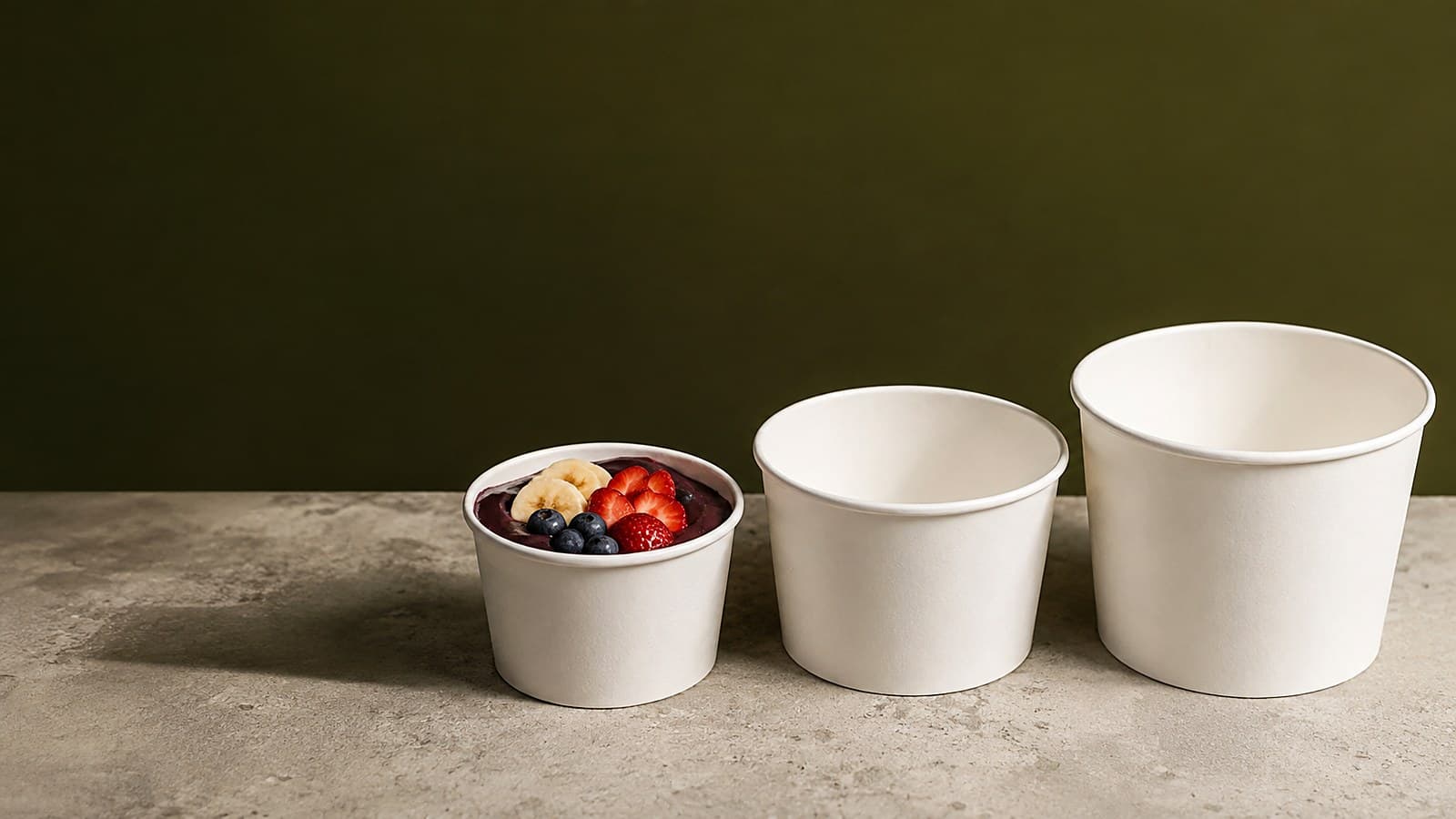 Três potes de papel branco em tamanhos crescentes sobre bancada de pedra, o menor servido com açaí, banana e frutas vermelhas, em fundo verde-oliva.